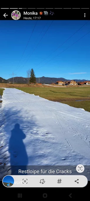 Eine einsame Schneeloipe aus Kunstschnee zwischen eher grünen Wiesen. Am rechten Rand stehen Häuser aus dem Landkreis Garmisch-Partenkirchen
