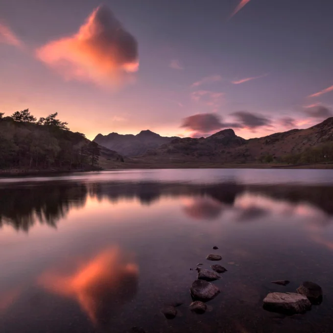 An image of a tarn with the water reflecting the clouds and hills surrounding it. With hints of pink and orange within the sky and clouds.