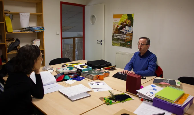 Sophie Blain, directrice de l'association Les Doigts Qui Rêvent est assise à une table en face de Bruno Gendron, président de la Fédération des Aveugles et Amblyopes de France. Il écrit sur une plage braille. Sur la table sont disposés plusieurs livres tactiles.