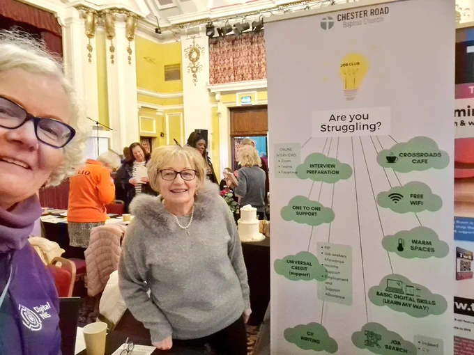 Two white women at an event either side of a table with a popup behind the table 