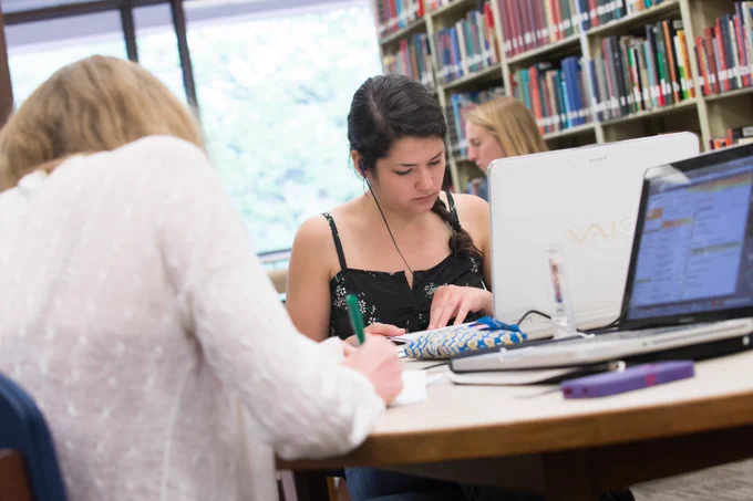Students writing and working on computers at library tables