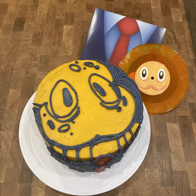  A photo of an Octodad layer cake with Octodad's charming face drawn on it with icing. The cake is posed next to the Octodad 7" vinyl and its packaging, which shows Octodad's suit.