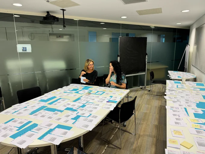 Our Technical Program Chairs, Corina Sas and Julie R. Williamson, are seated around a conference table that is covered with documents. They are engaged in a discussion regarding the organization of the technical program.