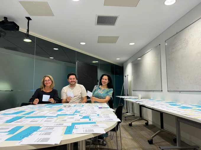 From left to right: Corina Sas, Kieran Waugh, and Julie R. Williamson are seated around a conference table filled with papers and documents. Each is holding a small white card. 