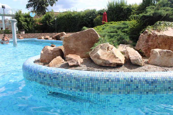 A bright blue pool, with a curved edge. At the side of the pool is a rockery filled with sand-coloured stones and green plants.