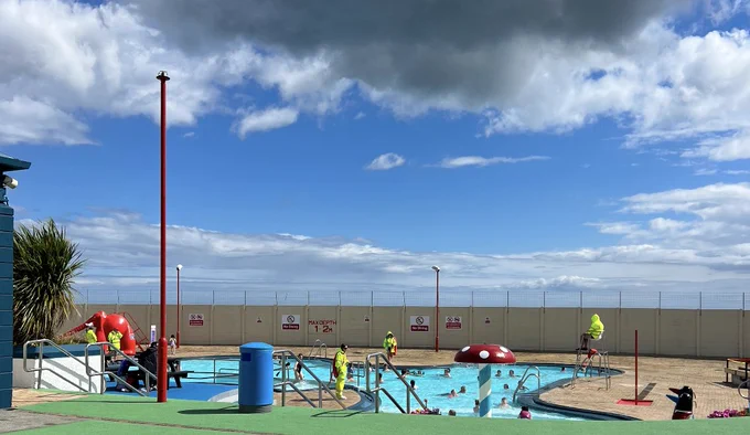 A curving fun pool under a blue and stormy sky. In the middle of the pol is a toadstool, painted bright red with white spots.
