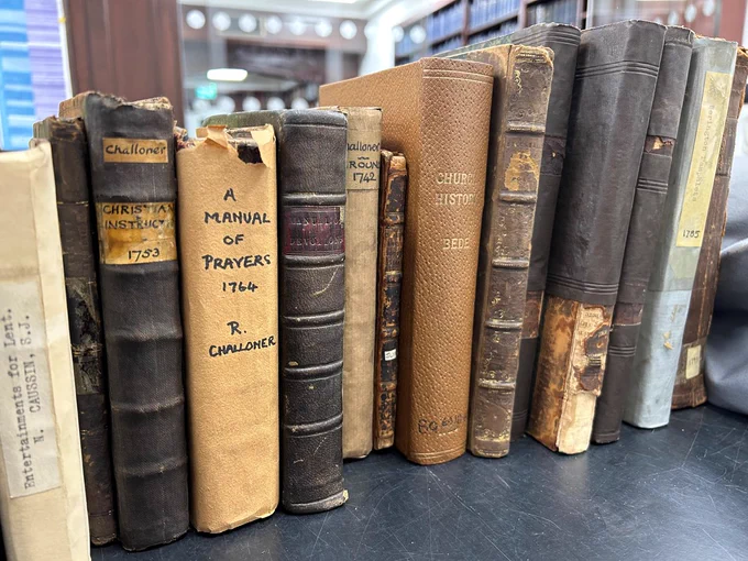 A row of about 14 books on a trolley, waiting to be catalogued.  Some have leather covers, some have had covers of brown paper and sellotape made.  All look a little tatty.