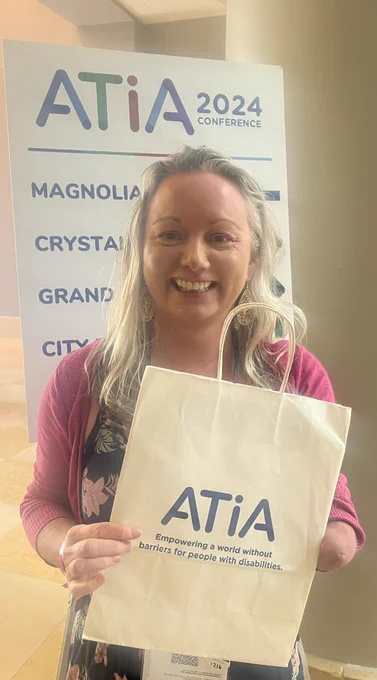 White woman with blonde hair, smiling & looking straight at camera. She has upper limb difference. Wearing a blue flowery jump suit & pink cardigan. She is holding a bag with ATiA logo in blue writing, & words “Empowering a world without barriers for people with disabilities”. She is standing infront of an ATIA sign at the conference.