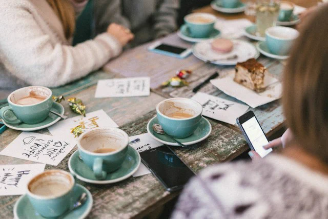 Multiple cups and saucers that are all the same style, size and colour on a narrow and long wooden table. The number of cups (that have different levles of cofee or tea left in them) indicates that it's a cafe in which people are sitting, talking and looking at their smartphones.