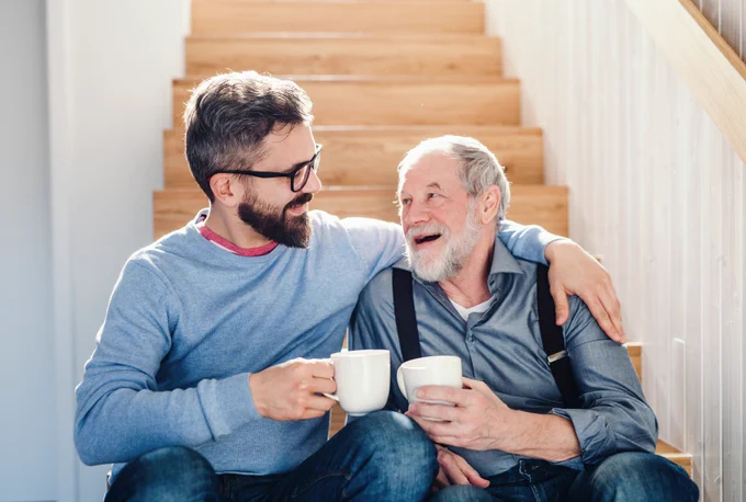 Two white men sit on a staircase holding cups of tea, smiling in conversation. The younger man has a beard and glasses, and his arm around the older man who wears a grey shirt with black suspenders.