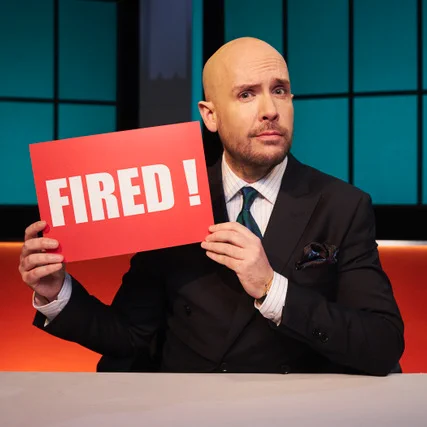 An image of Tom Allen in a black suit, sitting behind a desk, holding up a red sign with the word "FIRED!" written on it in white font.