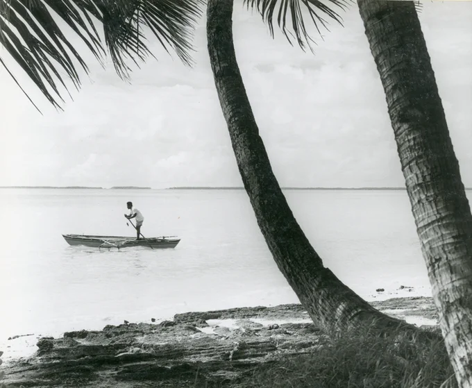 View of shoreline and ocean. Man on small boat paddles along. 