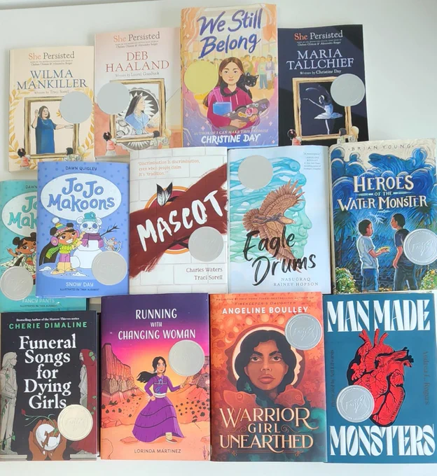 An overhead photograph of thirteen books laid out on a table. They all have colorful Native American-inspired artwork covers. Each book cover has a silver medal or seal, which signifies the book as a recipient of the 2024 American Indian Youth Literature Awards.