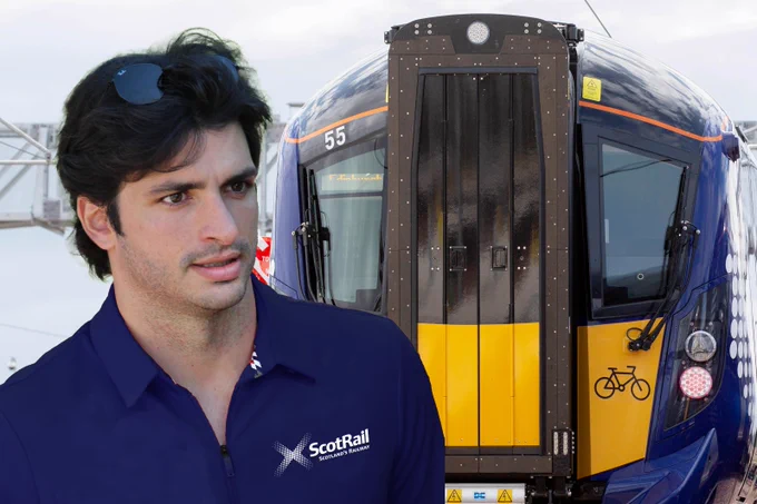 Formula One driver Carlos Sainz is stood beside a ScotRail train, number 55. He's wearing a blue ScotRail polo shirt. He looks confused,