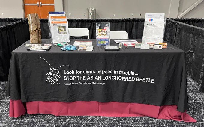 Photo of Asian longhorned beetle display table at the Tri-State Green Industry (OH). Tablecloth has an image of an Asian longhorned beetle and the words "Look for signs of trees in trouble... Stop the Asian longhorned beetle"
