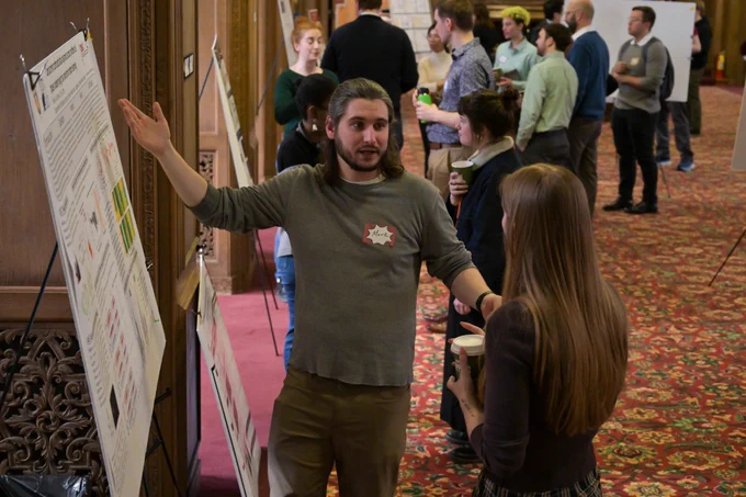 A man stands at a poster and tells a woman about his research.
