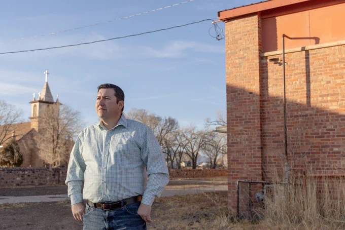 Armando Valdez stands looking out of frame to the left with a brick building and a church in the background