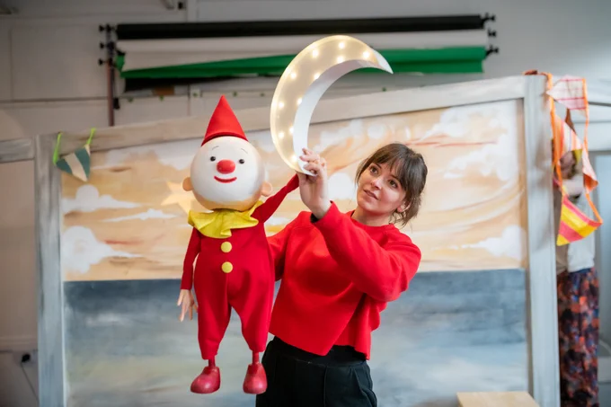 A clown puppet holding a light up moon as if it is floating in the sky. The actor operating the puppet is wearing a red toop and smiling.