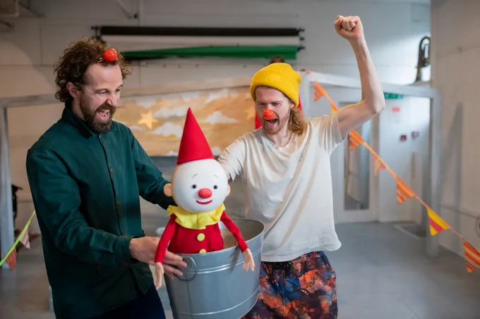 Two actors are holding a Clown puppet who is sailing in a tin bath. The actors are wearing red noses and cheering!