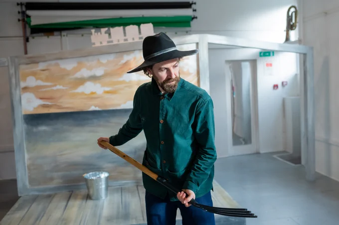 Actor standing alone, wearing a black, wide rimmed hat and holding a gardening fork. He is looking down and thoughtful.