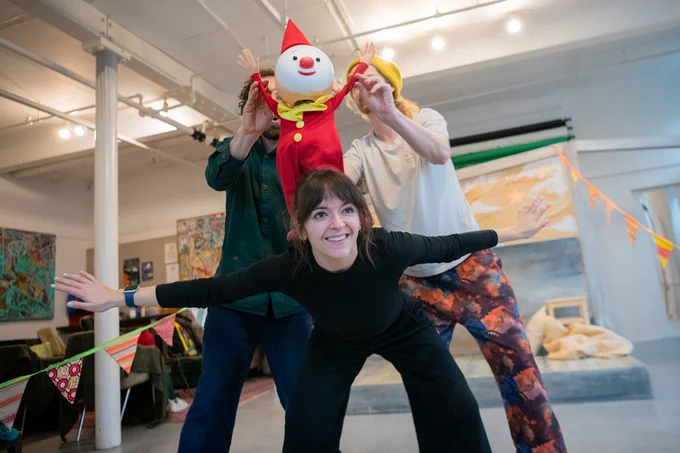 The Clown puppet is balancing on the back of one of the actors who is smiling. The puppet is being operated by two more actors. 
