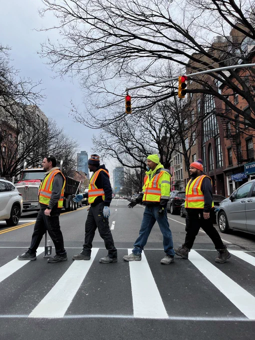 Four people in yellow and orange high visibility vests walk across a new crosswalk in the style of the Beatles abbey road cover
