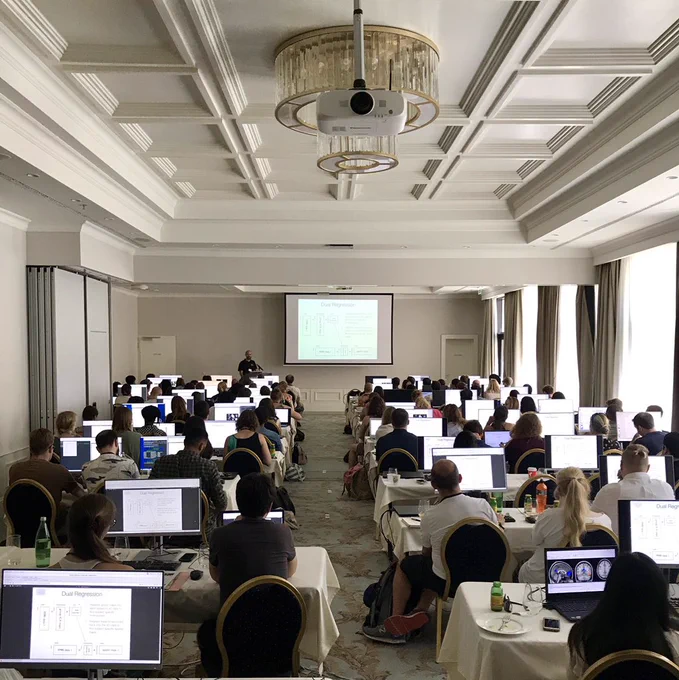 At the back of classroom facing a projector screen with slides. Several dozen students sit at desks facing the screen, with computer monitors lit. The walls and ceilings are white. In-person FSL Course 2019 in Croatia. Photo Credit: Rick Lange