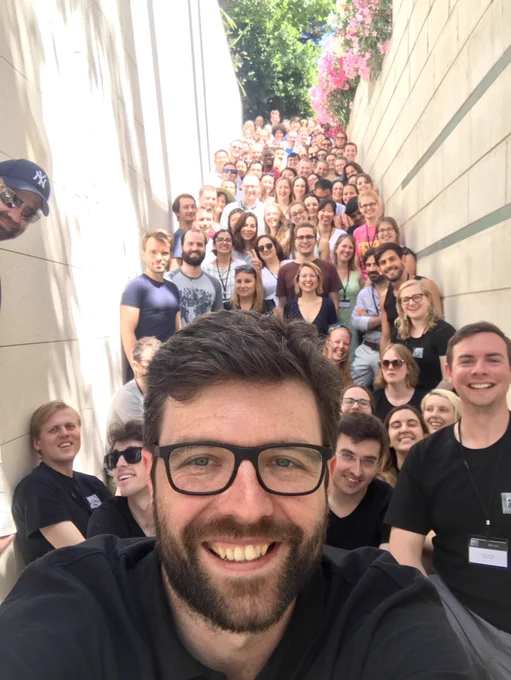 A white man with brunette short hair and beard, and glasses, smiles at the camera, taking a selfie with several dozen people behind him on a series of ascending stairs in a narrow outdoor stair corridor with light walls. In-person FSL Course 2019 in Croatia. Photo Credit: Rick Lange