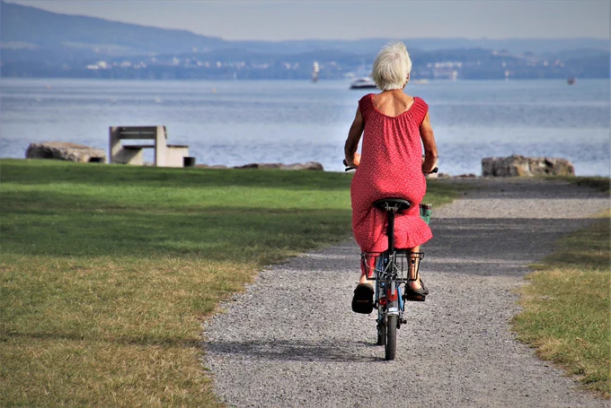 A photograph of a woman in a red dress riding a bicycle towards a lake on a gravel path surrounded by grass