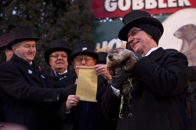 A group of older men in suits and top hats stand on a stage. The man in the center holds a scroll of paper. The man on the right smiles and holds a groundhog. 