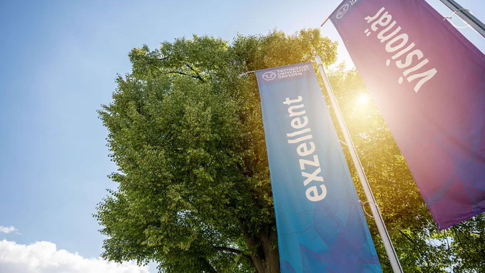Zwei Flaggen mit den Aufschriften "exzellent" und "visionär"; im Hintergrund ein Baum und blauer Himmel