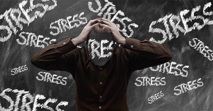 Image from Pixabay.
A person stood in front of a blackboard holding their head with their hands. On the black board the word 'stress' is written/projected all over, including across the person's face.