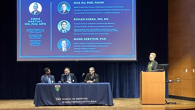 A photo of Rohan Khera, Hua Xu and Mark Gerstein seated on stage with a tablecloth that says "Yale Biomedical Informatics & Data Science". Annie Hartley stands at the lectern, moderating.