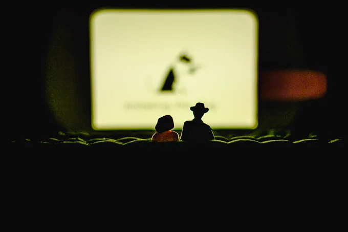 📸by Ed Waring. A model cinema interior. Two tiny figures seated, one wears a hat. They are watching a cinema screen displaying an out-of-focus image of the ITD logo. From installation Seeing in the Dark