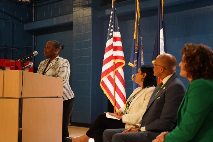 Parent speaking at a podium at PS 958 for a Special Education Announcement. Chancellor Banks and N Y C Public Schools Administration are seated by the podium. 