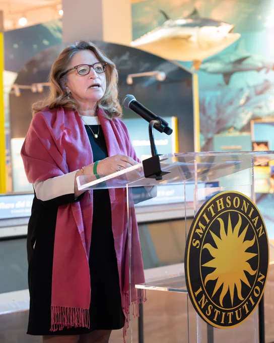 Ellen Stofan, Under Secretary for Science and Research at the Smithsonian, stands at a lucite podium with the Smithsonian logo on the front. She's speaking in the Ocean Hall at the Smithsonian National Museum of Natural History. She has shoulder-length brown hair, black glasses, a black dress, and a pink shawl over her shoulders. 