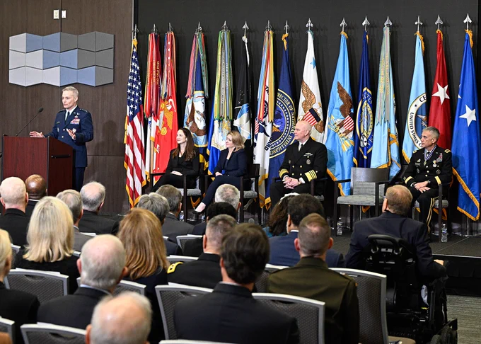 A man in uniform stand behind a podium speaking to an audience, while a group of men and women sit on stage in front of a row of colorful flags.