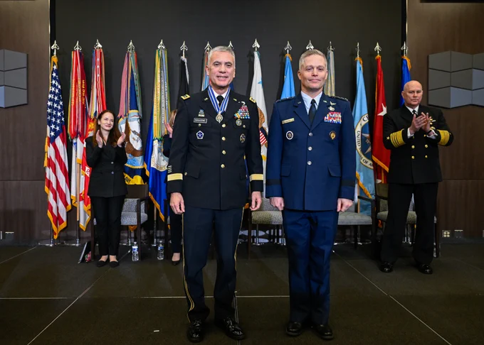 Two men in uniform stand next to each other smiling, in front of a row of colorful flags.