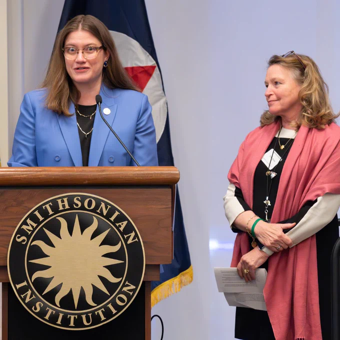 Sarah Kapnick, Ph.D., NOAA Chief Scientist speaks at a podium with the Smithsonian logo on the front. She has shoulder length brown hair, brown glasses, and is wearing a light blue suit with a black top. Ellen Stofan, Under Secretary for Science and Research at the Smithsonian, stands next to her listening. Ellen is wearing a black dress with a pink shawl. She has brown hair and her black glasses are on top of her head. 