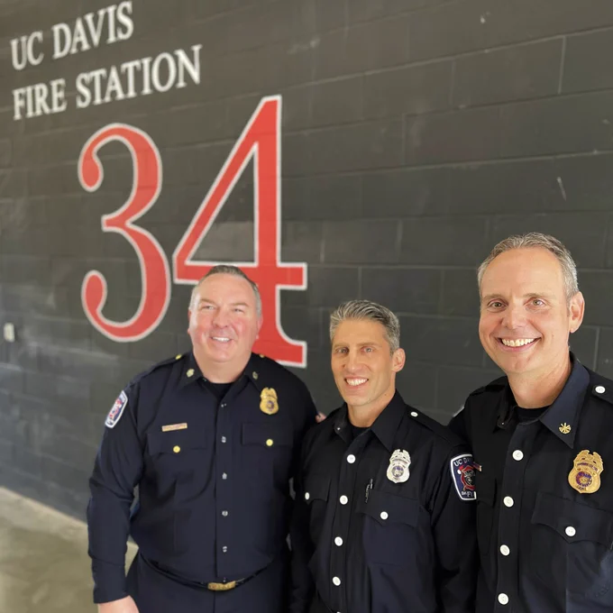 Three men in black uniform smile in front of wall that says ‘UC Davis Fire Station 34’.