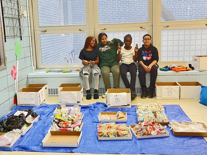 Four 5th grade students from Cafeteria Culture partner school, PS/MS 34 FDR in Lower Manhattan, sit together behind a blue tarp on the floor with the single-use plastic from one lunch period, sorted into labeled cardboard trays.  Labeled cardboard boxes next to the tarp were used for cafeteria sorting.
