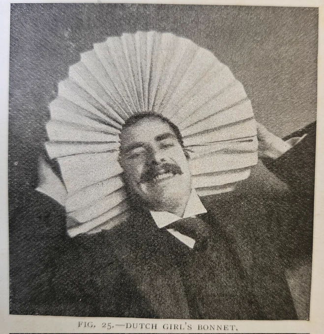 A black and white Victorian photograph of a smiling gentleman. He is wearing a suit and tie, and he is holding a large folded piece of card over his head in the shape of a bonnet. The caption beneath the photograph reads: "Fig. 25. - Dutch Girl's Bonnet."