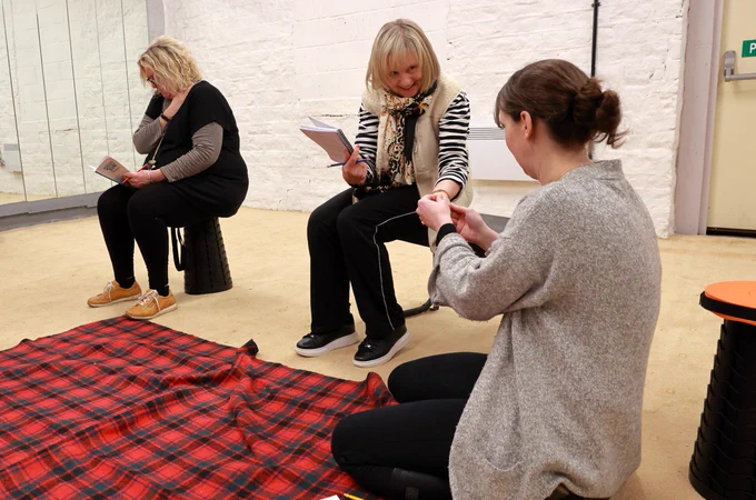 Three actresses rehearse Sheila's Island - at this point, sitting around a rug and rationing out their only sausage - ready for Longwood Mechanics Hall, 13-16 March.
