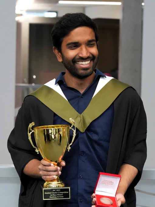 Smiling man in graduation robes holding trophy cup and boxed medal
