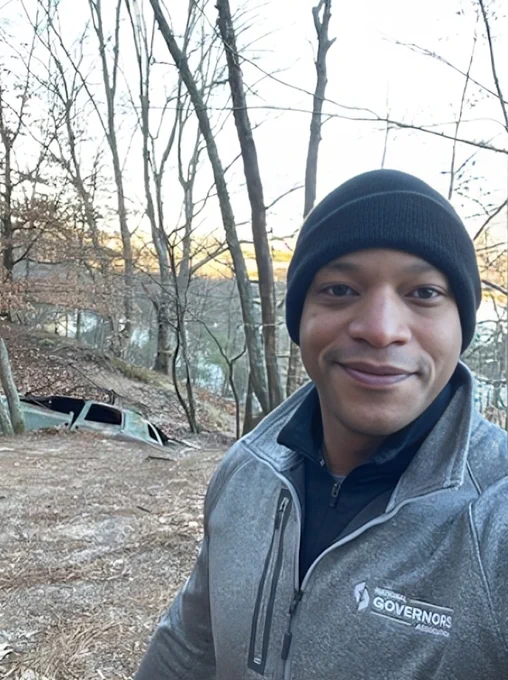 Governor Moore taking selfie in Tuckahoe State Park, with buried vehicle in background