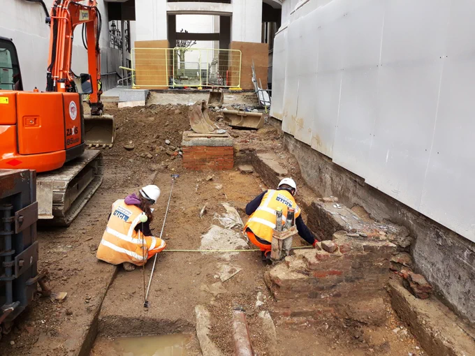 A small excavation site with two archaeologists at work with their backs to us, wearing hard hats and high vis clothing. A few partial walls are emerging from the ground.