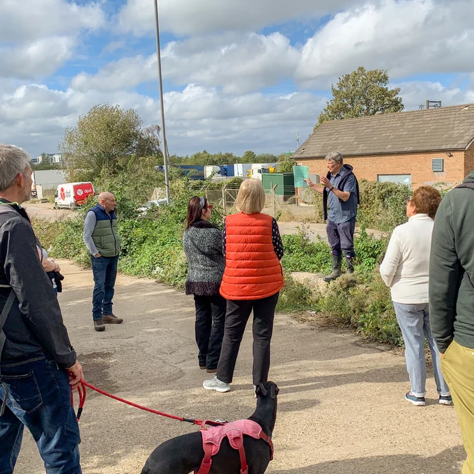 A person stands on a small green verge, talking to a group of people who are gathered listening to them speak. 
