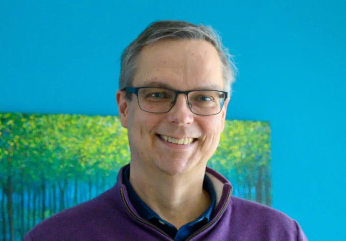Professor David Stephens, smiling, wearing glasses and purple top, against blue background.