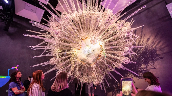 Ein Foto zeigt die Installation Noosphere im Futurium Lab. In dessen Zentrum leuchtet es. Davon ab ragen strahlenförmig Metallteile in den Raum. Drum herum stehen mehrere Personen und eine Person macht ein Foto mit dem Smartphone. Foto Credit: Jan Windszus.