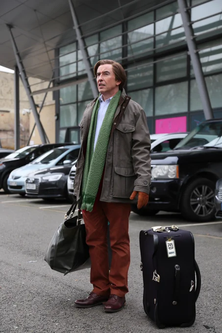 Alan Partridge stands with a suitcase and bag in hand, wearing a coat and scarf.